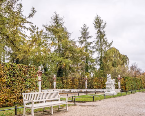 White benches and classical statues line a gravel path in a landscaped park with tall trees and trimmed hedges on an overcast day.