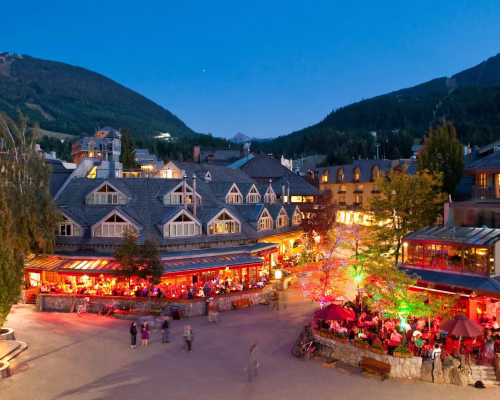 An evening view of a lively village square with lit restaurants, outdoor seating, people walking, and mountains in the background.