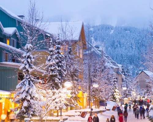Snow-covered street in a mountain village with colorful buildings, pine trees, and people walking in winter clothing during dusk.