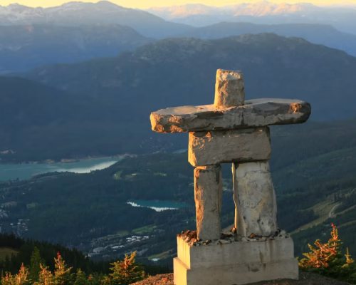Stone inuksuk statue stands on a mountain overlooking a green valley, forests, lakes, and distant mountain ranges under soft sunlight.