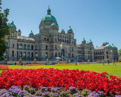A large historic building with a green dome roof, surrounded by manicured lawns and vibrant flower beds under a clear blue sky.