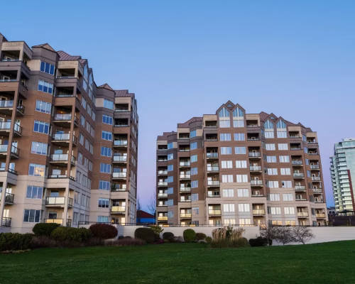 Two mid-rise apartment buildings with numerous balconies stand next to a grassy area, with taller buildings visible in the background under a clear sky.