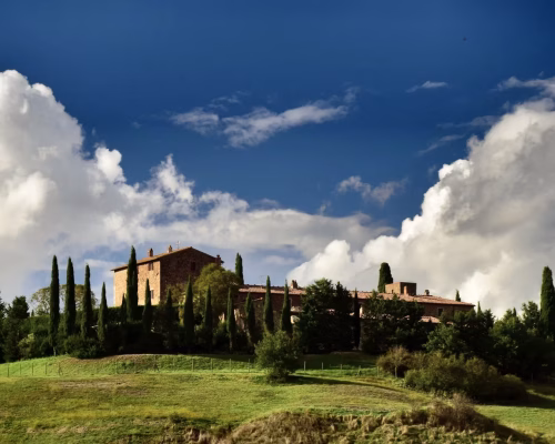 A rustic stone building surrounded by tall cypress trees sits atop a grassy hill under a blue sky with large white clouds.