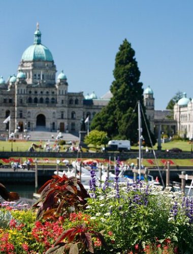 The image shows the British Columbia Parliament Buildings behind a harbor with boats, colorful flowers in the foreground, and people on the lawn under a clear blue sky.