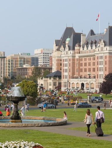 People walk and gather near a fountain in a park, with historic buildings and a large hotel in the background under a clear sky.