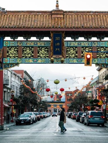 A large, ornate Chinese gate spans a busy city street with cars parked along both sides and pedestrians crossing underneath. Red lanterns hang above the street.