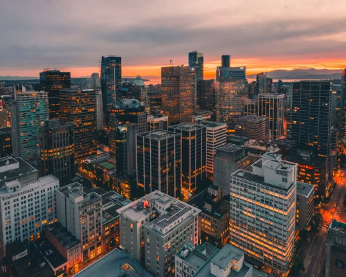 A city skyline at sunset with illuminated office buildings and streets, under a cloudy sky with orange hues on the horizon.