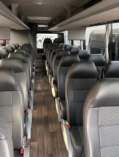 Interior view of an empty passenger bus with rows of black leather seats, overhead storage, and large windows on both sides.