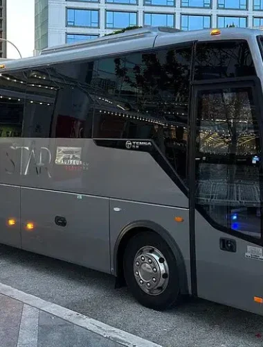 A gray mid-sized coach bus is parked on a city street near glass buildings on a sunny day.