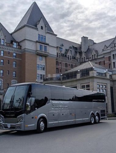 A large gray tour bus is parked in front of a historic, castle-like building on a cloudy day.