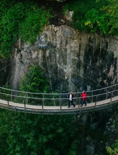 Two people walk on a curved suspension bridge over a forested gorge with rocky cliffs and dense greenery.