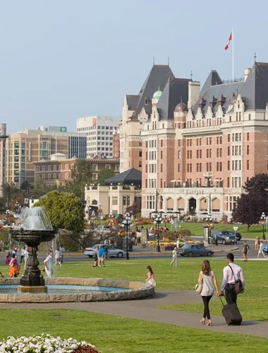 People walk and relax on a grassy area with a fountain in front of a large historic hotel building, with city buildings in the background.