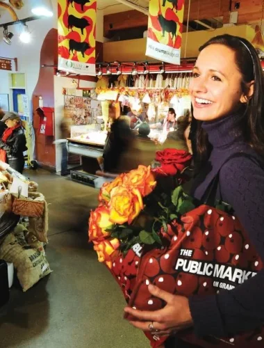 A woman carrying a bouquet of roses and a polka-dot shopping bag stands inside a busy public market filled with food stalls and shoppers.