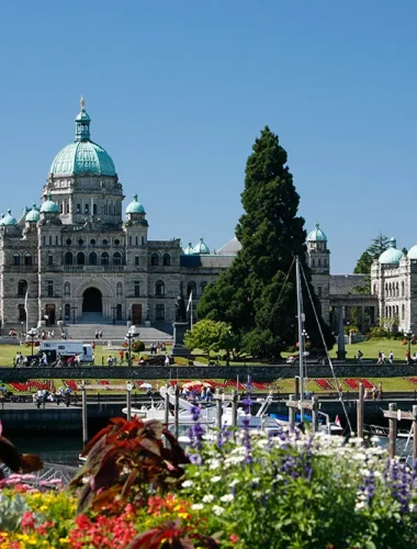 The exterior of the British Columbia Parliament Buildings in Victoria, Canada, viewed from across the harbor with flowers and boats in the foreground.
