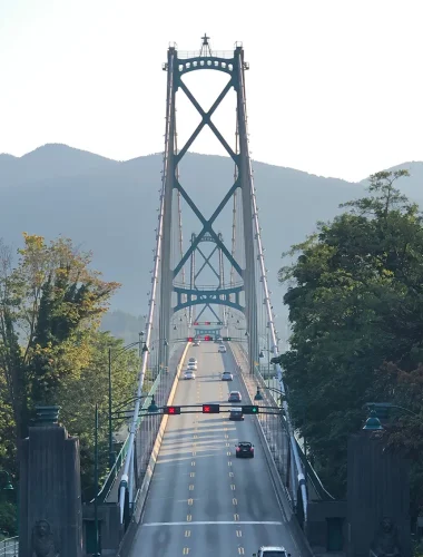 A suspension bridge with cars crossing, surrounded by trees and hills in the background on a clear day.
