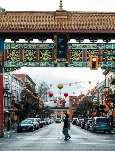 A person crosses the street under a traditional Chinese archway in a busy urban Chinatown, with cars parked along the road and red lanterns hanging above.