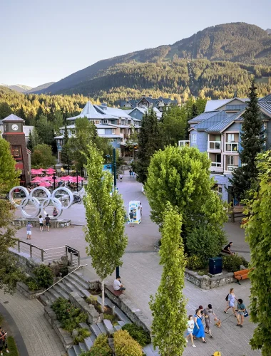 An outdoor pedestrian plaza with people walking and sitting, surrounded by trees, modern buildings, and mountains in the background.