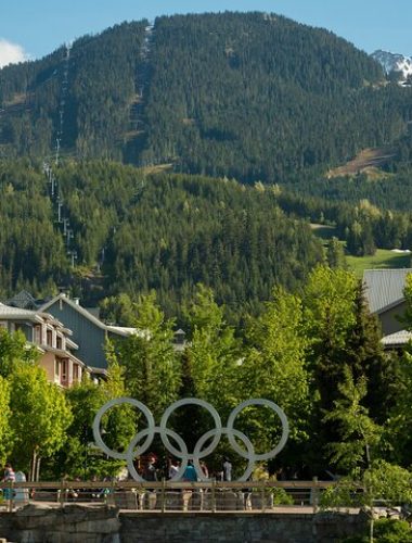 The Olympic rings monument stands in a landscaped area with buildings and forested mountains in the background on a sunny day, making it a popular stop for Sightseeing Tours Whistler.