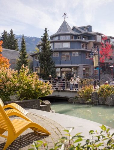 Yellow lounge chairs face a small stream and arched bridge, offering a relaxing spot to unwind after Sightseeing Tours Whistler, with a multi-story building and autumn trees in the background under a clear sky.