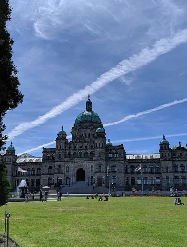 A large historic building with green domes stands under a blue sky with white streaked clouds, surrounded by a grassy lawn and scattered people.