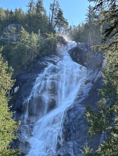 Water cascades down a rocky cliff surrounded by dense green trees under a clear blue sky—a perfect spot for Sightseeing Tours Whistler.