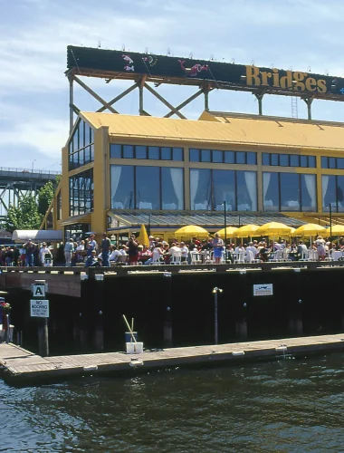 A large yellow waterfront restaurant with outdoor seating under yellow umbrellas, crowded with people, next to a dock and water, with a bridge visible in the background.