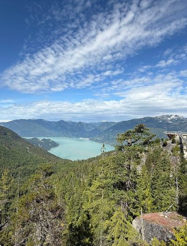 A scenic view of a forested valley with a turquoise lake, surrounded by mountains under a partly cloudy sky—perfect for sightseeing tours Whistler has to offer.