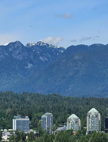 A cityscape with several mid-rise buildings surrounded by dense trees, set against a backdrop of forested mountains under a partly cloudy sky.
