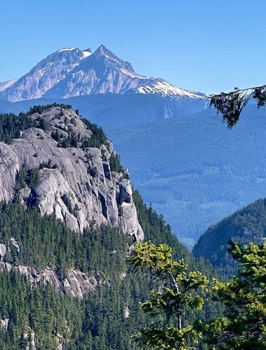 A rocky mountain peak rises above a forested landscape, with a snow-capped mountain in the background under a clear blue sky—the perfect scene for unforgettable Sightseeing Tours Whistler.