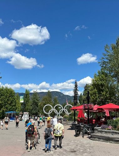 People walk through a plaza with the Olympic rings sculpture, surrounded by trees, outdoor seating with red umbrellas, and a clock tower under a sunny blue sky—a perfect spot for Sightseeing Tours Whistler.