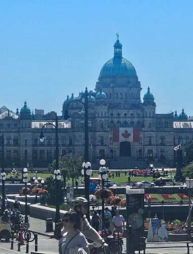 The British Columbia Parliament Buildings in Victoria, Canada, with a large Canadian flag displayed on the front and people gathered in the foreground on a sunny day.