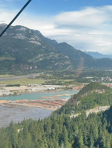 A wide view of a river with log booms, industrial buildings, and forested mountains under a partly cloudy sky—perfect for those on Sightseeing Tours Whistler seeking breathtaking natural and industrial scenery.