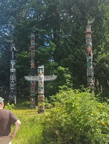 Several people observe a group of tall, colorful totem poles standing among trees and greenery in an outdoor park setting.