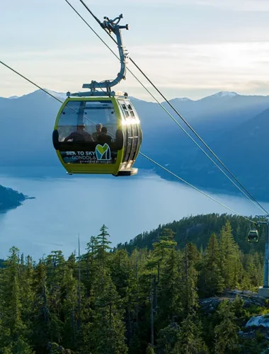 A gondola cable car travels above a forested mountain overlooking a large body of water, with mountains visible in the background.