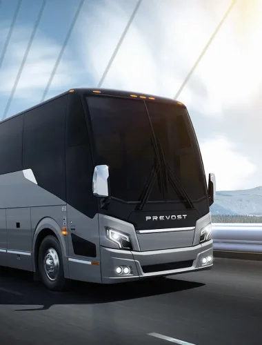 A large, modern Prevost coach bus drives on a bridge with mountains and a bright sky in the background.