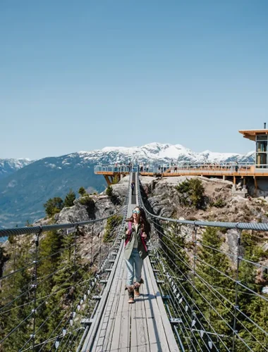 Person walking across a suspension bridge with snow-capped mountains and a modern building in the background under a clear blue sky.