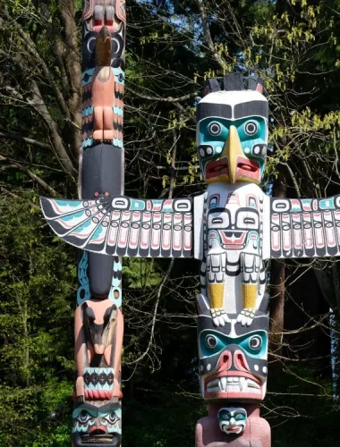 A large, colorful totem pole with bird and animal carvings stands outdoors in front of green trees.