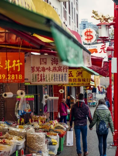 Two people walk through a busy street market lined with colorful awnings, food stalls, and signs with Chinese characters in an urban Chinatown area.
