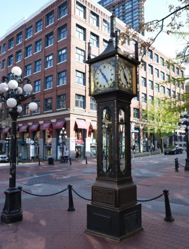 A steam clock stands on a brick-paved street corner in front of a historic red brick building with shops and large windows.